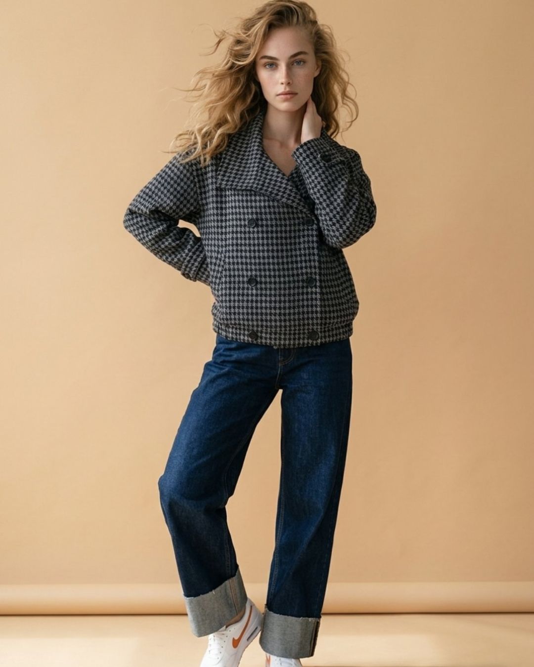 Grey Houndstooth Wool Coat A woman modeling a black and white gingham double-breasted blazer with dark blue jeans and white sneakers, standing against a solid beige studio backdrop.