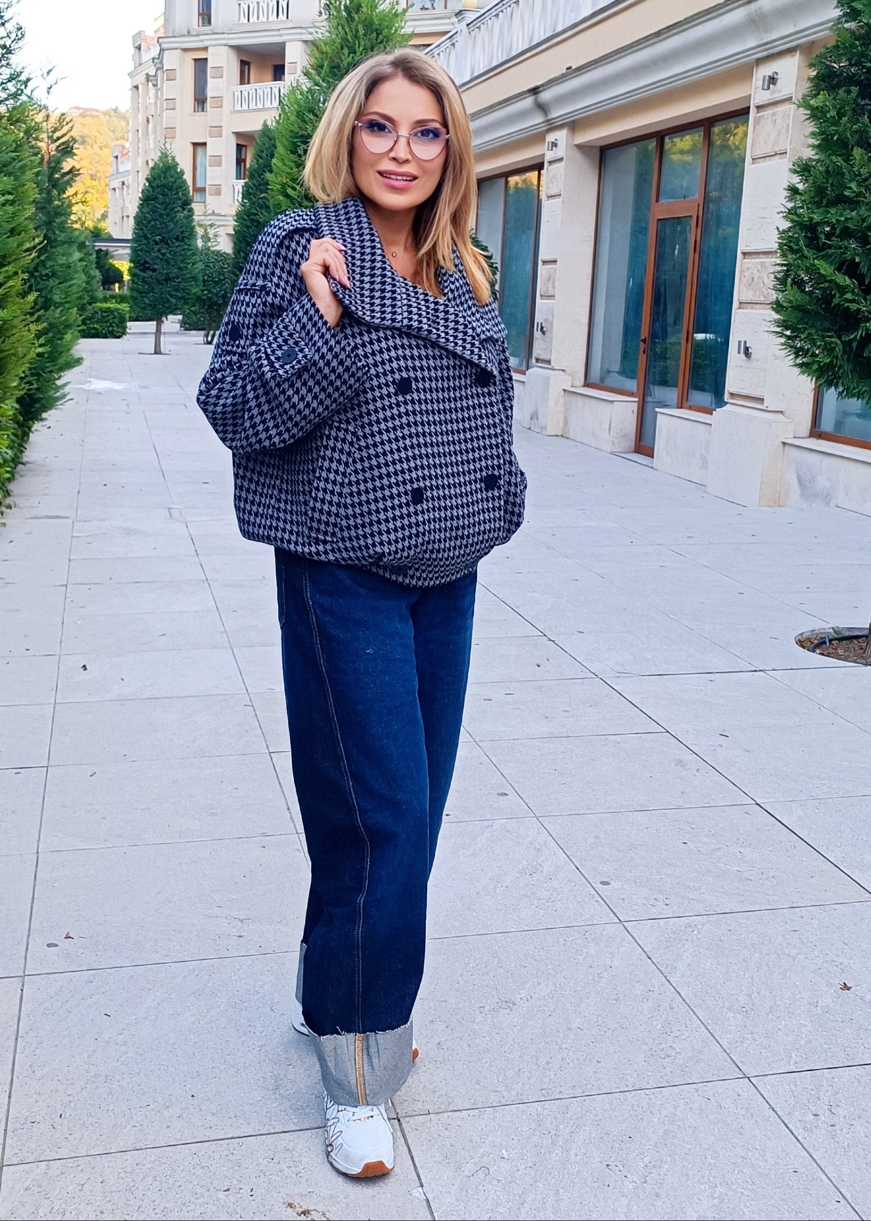 Grey Houndstooth Wool Coat A woman wearing a navy and beige houndstooth blazer, dark blue jeans, and white sneakers stands on a paved outdoor walkway in front of a building.