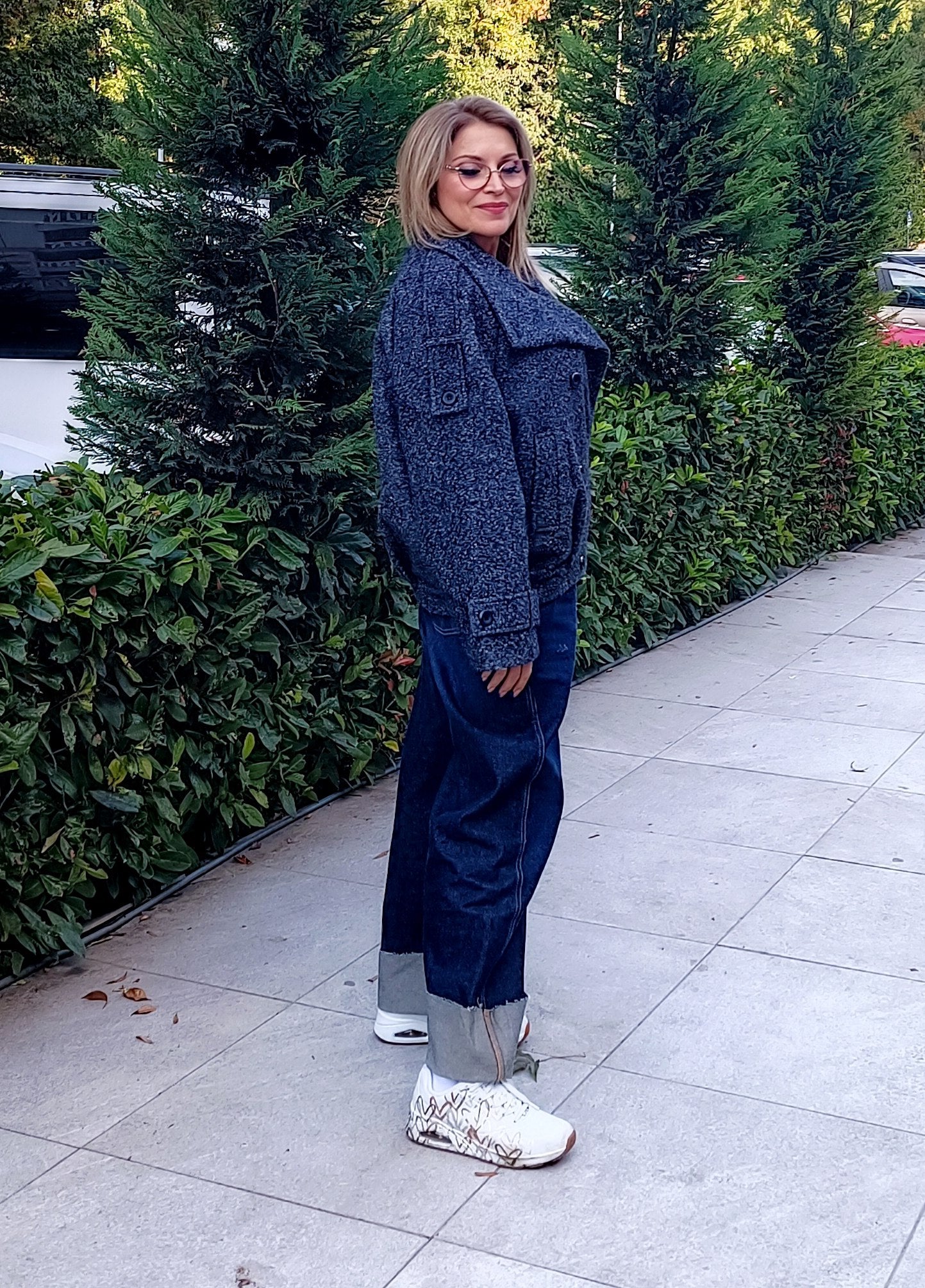 Grey Bouclé Wool Coat A woman wearing a dark blue textured knit jacket, dark blue wide-leg jeans, and white sneakers stands on a paved sidewalk outdoors with green hedges in the background.
