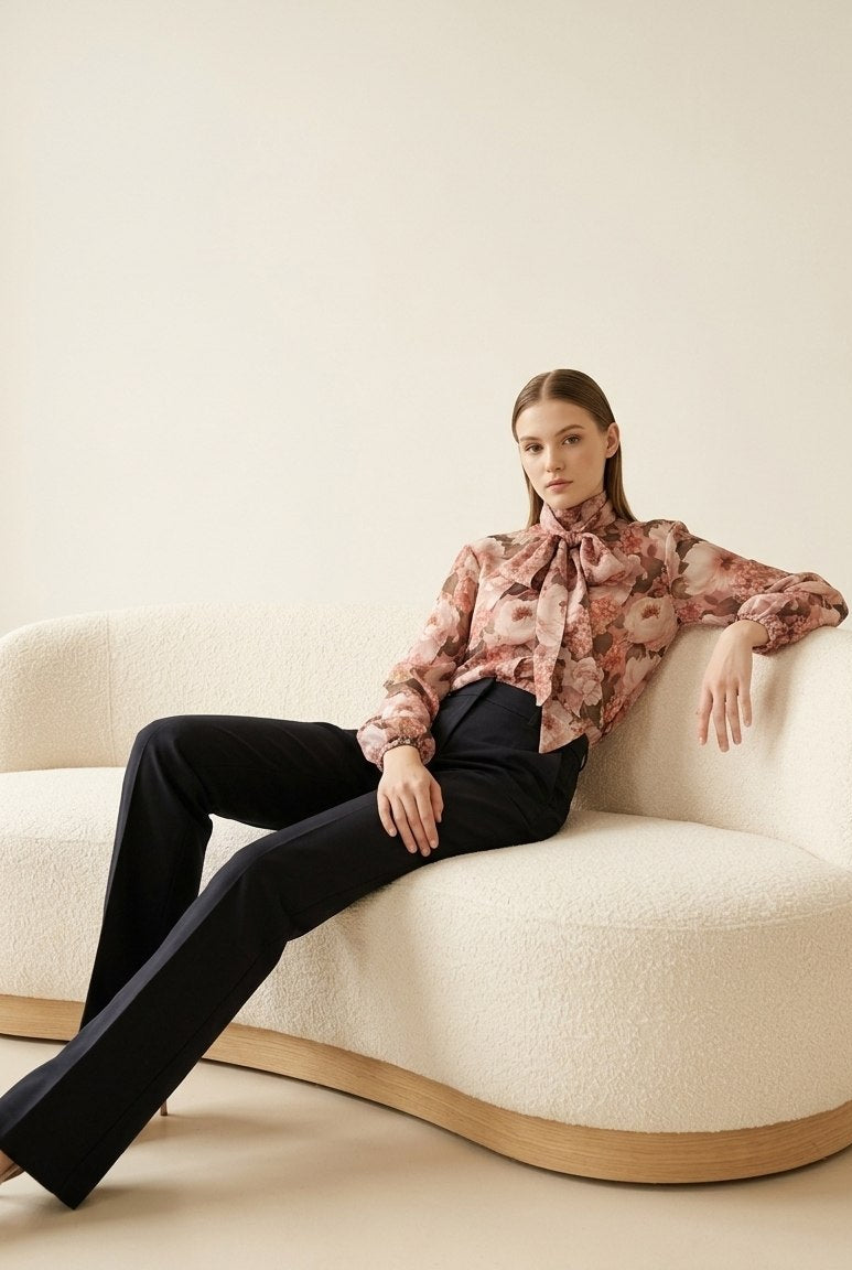 Silk Floral Blouse with Long Scarf Tie A woman sits on a curved cream boucle sofa wearing a long-sleeve floral blouse with a high neck and bow detail, paired with black trousers.
