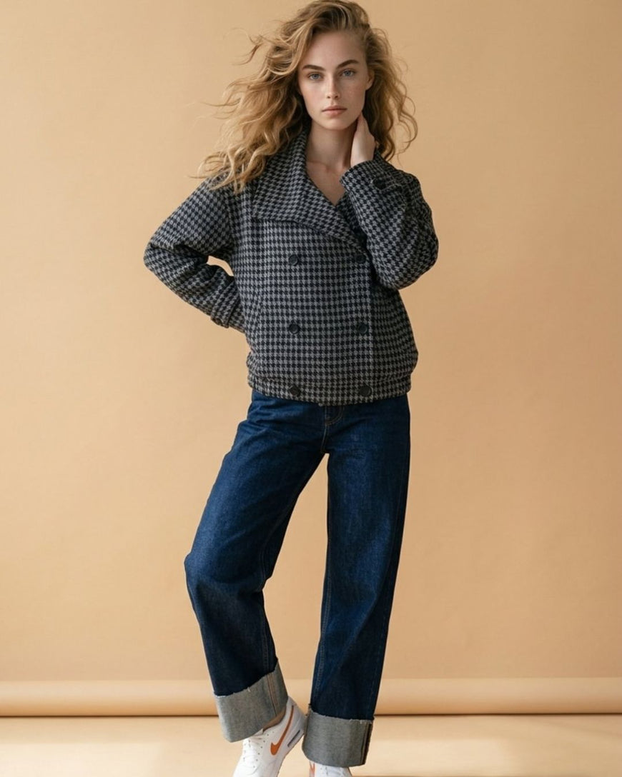 A woman modeling a black and white gingham double-breasted blazer with dark blue jeans and white sneakers, standing against a solid beige studio backdrop.