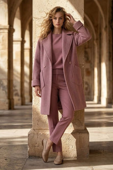 A woman stands in a sunlit stone arcade wearing a coordinated dusty pink outfit consisting of a long coat, top, and trousers, with beige flats.