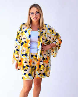 A woman modeling a matching two-piece lounge set featuring a yellow, orange, and navy floral kimono jacket and shorts over a light blue top, set against a plain white studio background.