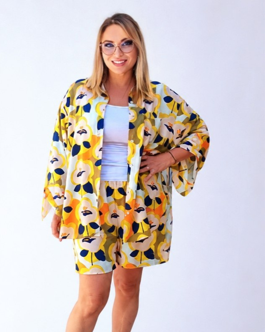 A woman modeling a matching two-piece lounge set featuring a yellow, orange, and navy floral kimono jacket and shorts over a light blue top, set against a plain white studio background.