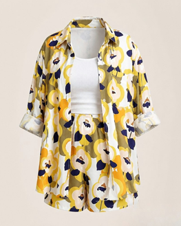 A women's two-piece matching set featuring a short-sleeve button-up shirt and shorts in a bold yellow, navy blue, and white floral pattern, styled over a white ribbed tank top on a mannequin against a solid beige background.