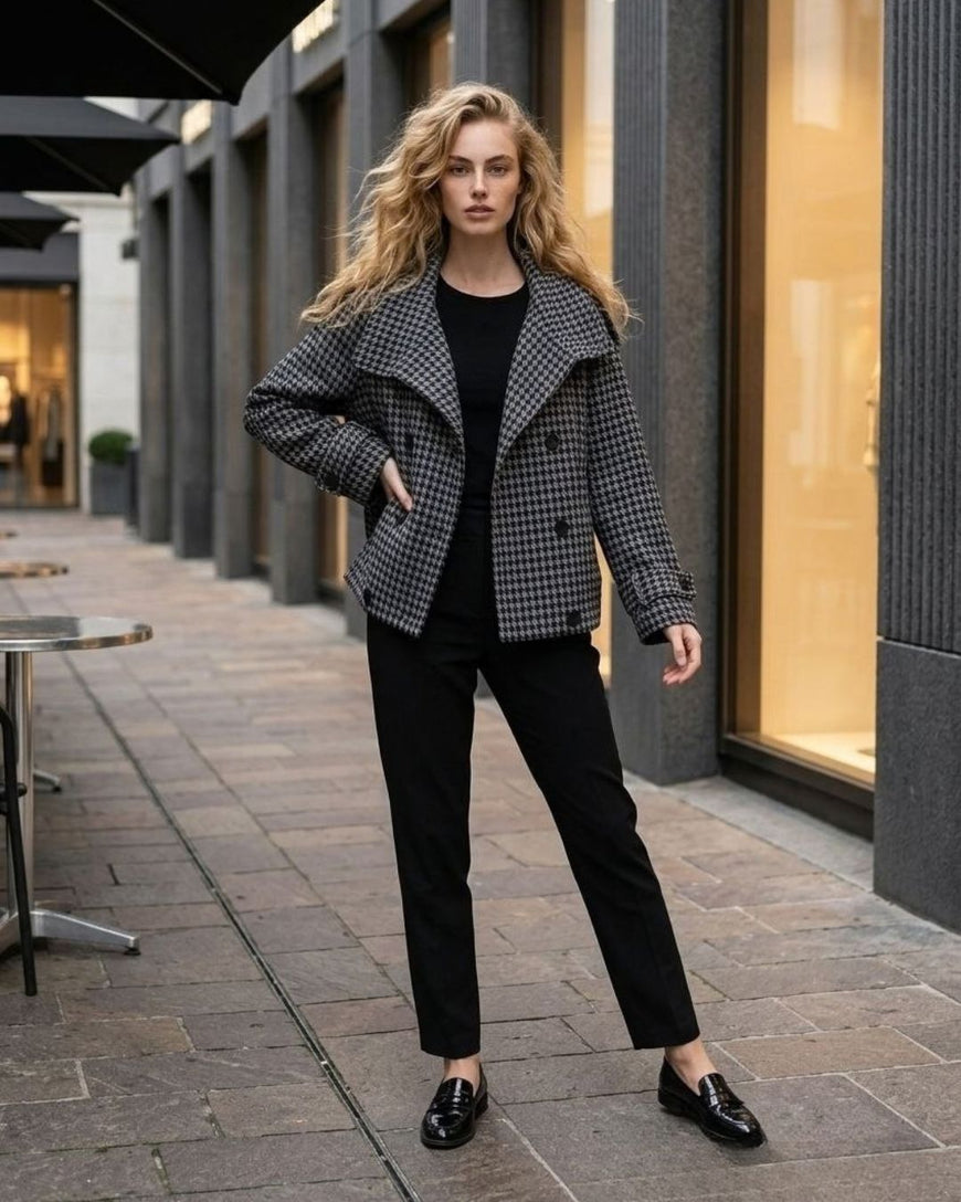 A woman stands on a city sidewalk wearing a black-and-white houndstooth blazer over a black top and black trousers, with black loafers. Storefront windows and outdoor seating are visible in the background.