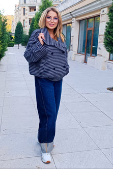 A woman wearing a navy and beige houndstooth blazer, dark blue jeans, and white sneakers stands on a paved outdoor walkway in front of a building.