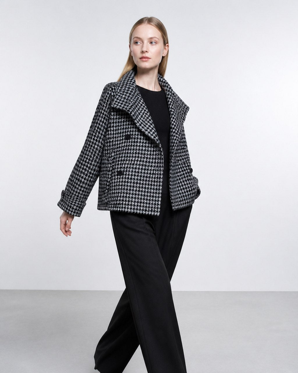 A woman modeling a black and white houndstooth blazer over a black top and black wide-leg trousers, walking against a light gray studio background.
