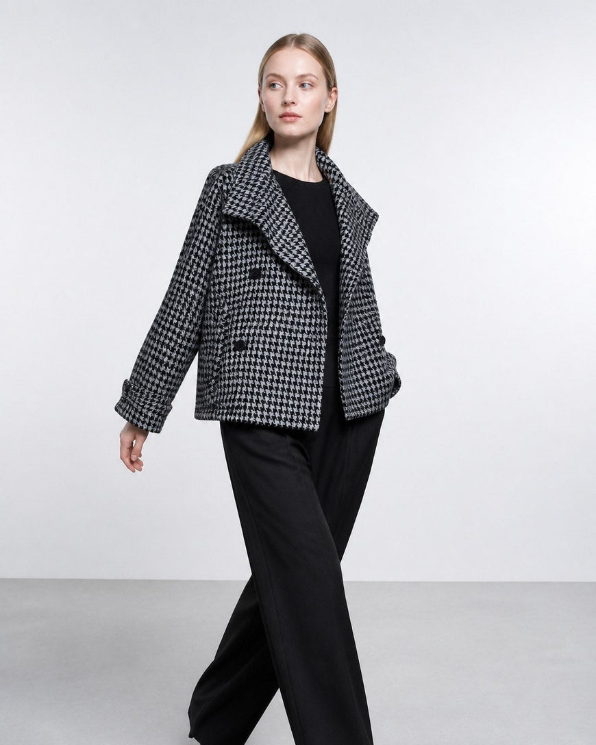 A woman modeling a black and white houndstooth blazer over a black top and black wide-leg trousers, walking against a light gray studio background.