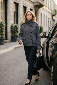 A woman walking on a city street wearing a black and gray houndstooth double-breasted turtleneck jacket, black trousers, and black heels, while carrying a black handbag.
