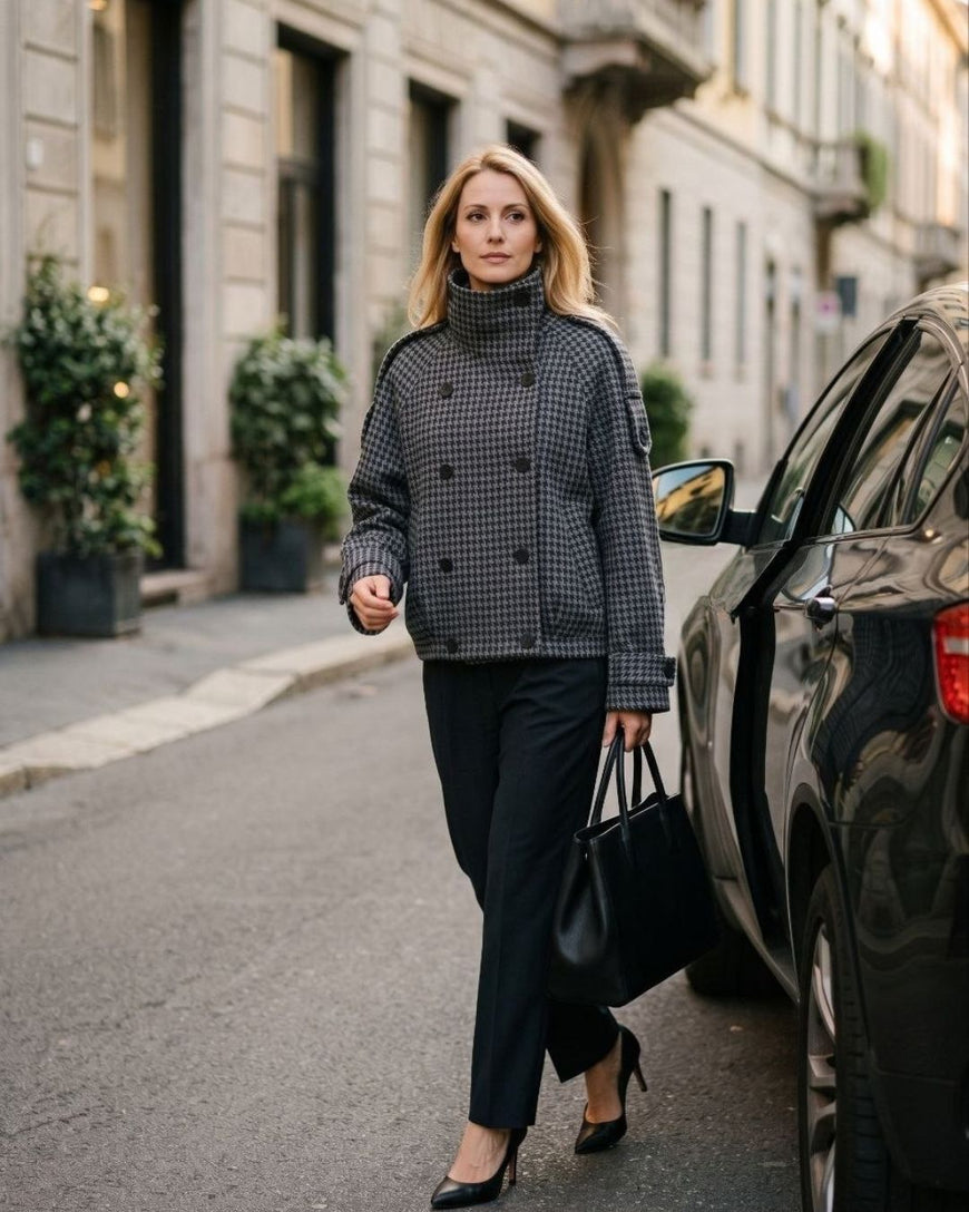 A woman walking on a city street wearing a black and gray houndstooth double-breasted turtleneck jacket, black trousers, and black heels, while carrying a black handbag.
