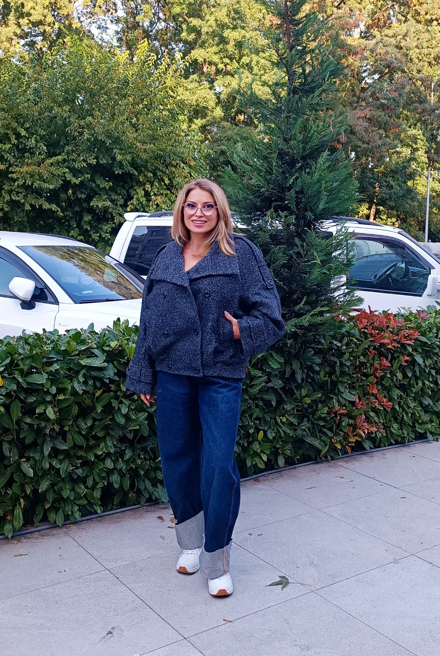 A woman wearing a dark gray bouclé jacket, blue jeans, and white ankle boots stands on a sidewalk outdoors in front of a hedge and parked cars.