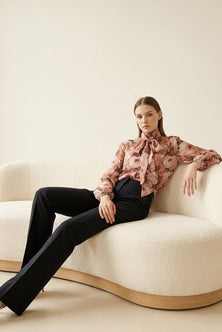 A woman sits on a curved cream boucle sofa wearing a long-sleeve floral blouse with a high neck and bow detail, paired with black trousers. 