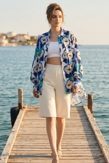 A woman walks on a wooden dock by the sea wearing a blue, white, and beige patterned button-up shirt over a white crop top and cream shorts.